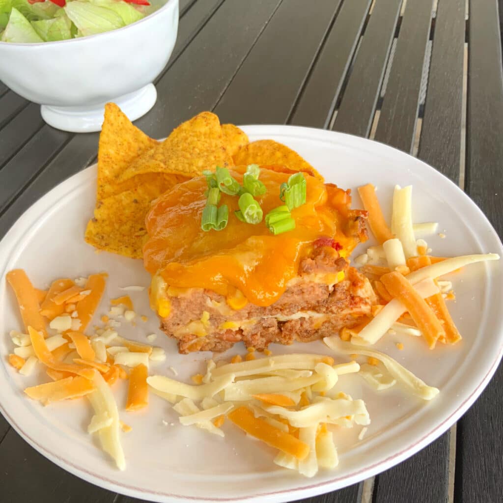 mexican lasagna on a round white plate with shredded cheese laying around it and a salad in the background.
