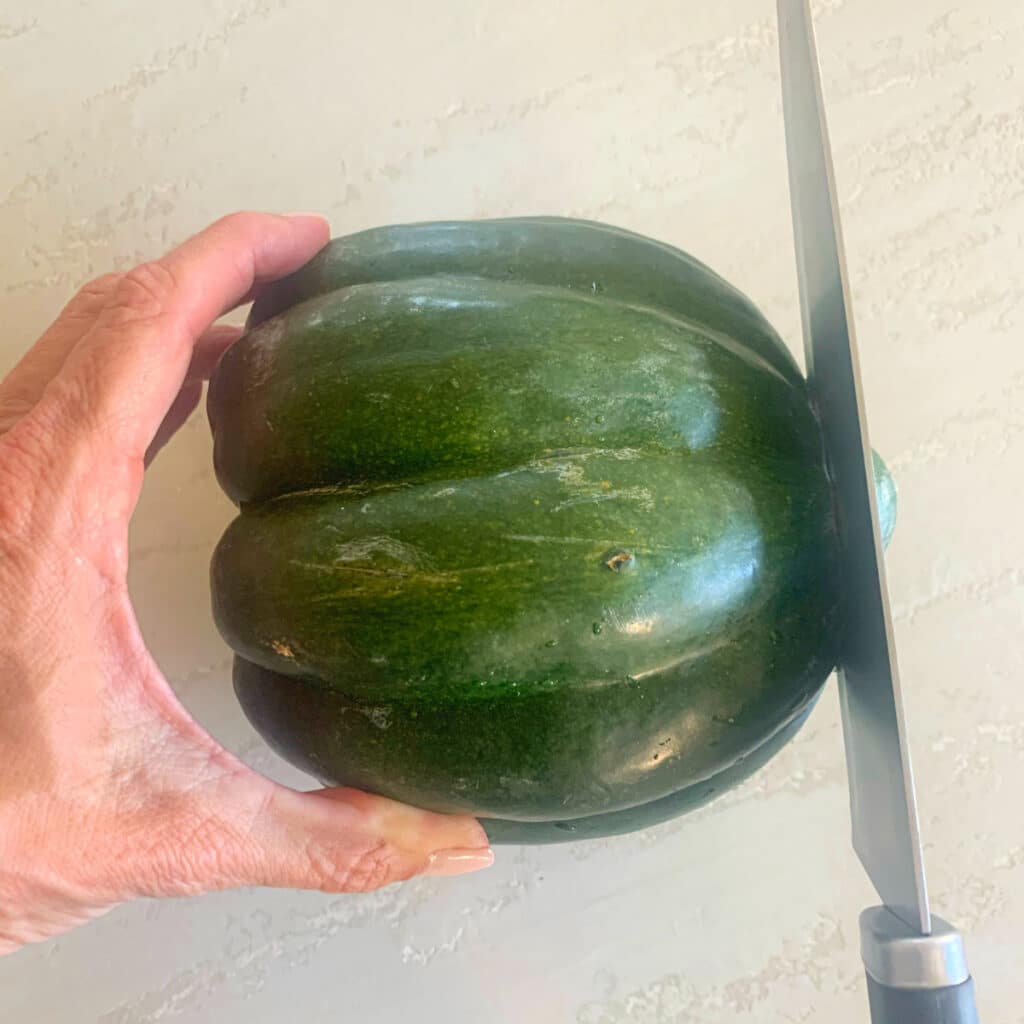 Whole acorn squash being held by a hand with a knife about to slice off the end