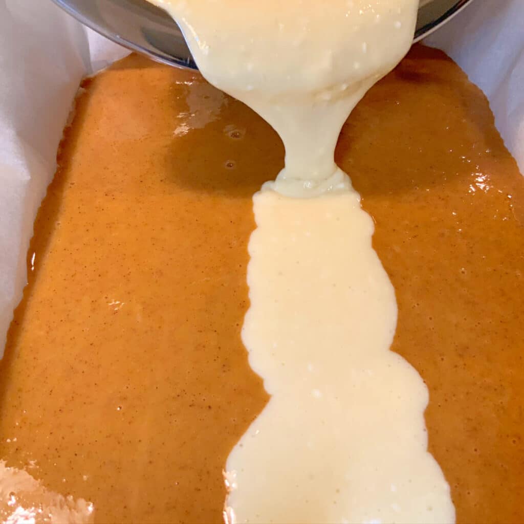 pumpkin layer of batter in a slow cooker lined with parchment paper and the cheesecake layer is being added as a white strip down the middle