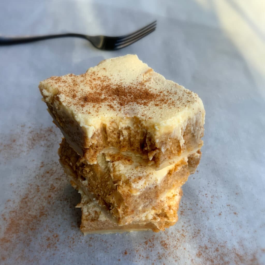 a stack of pumpkin cheesecake bars sprinkled with cinnamon. a bite is taken out of the top most bar.