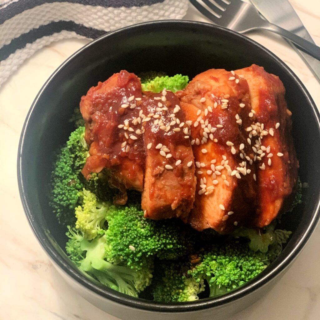 sliced chicken placed on lightly steamed broccoli in a round black bowl topped with sesame seeds