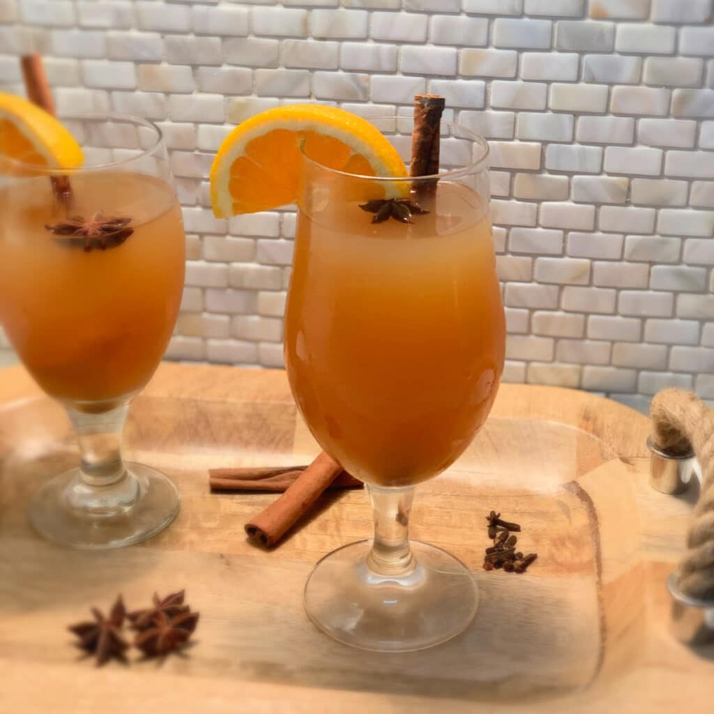 two glasses of hot spiked apple cider in a glass with garnish on a wooden tray with a rope handle