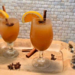 two glasses of hot spiked apple cider in a glass with garnish on a wooden tray with a rope handle