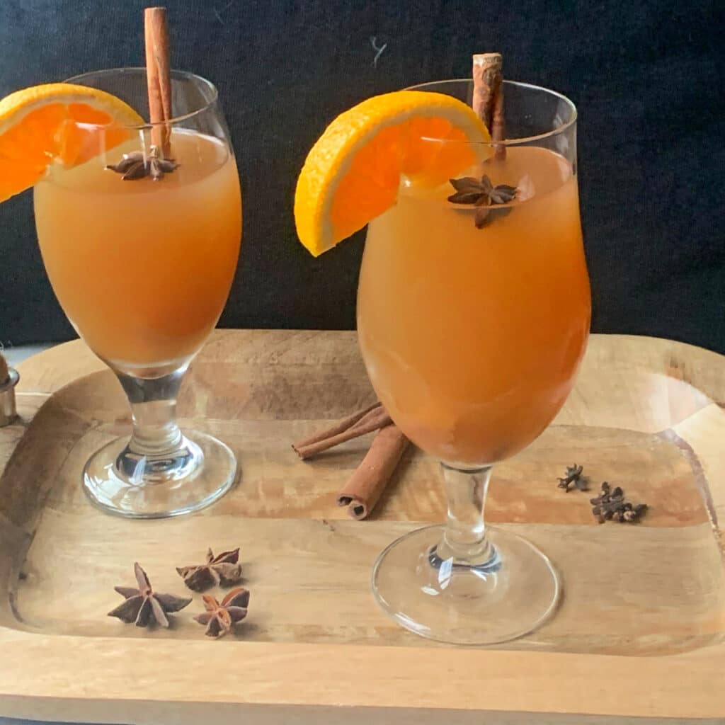 close up of two glasses of hot apple cider with star anise floating on top with garnish on a wood tray with a black background