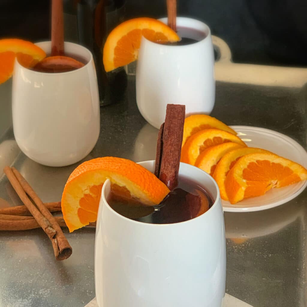 close up of three glasses containing mulled wine with a plate of sliced oranges and cinnamon sticks