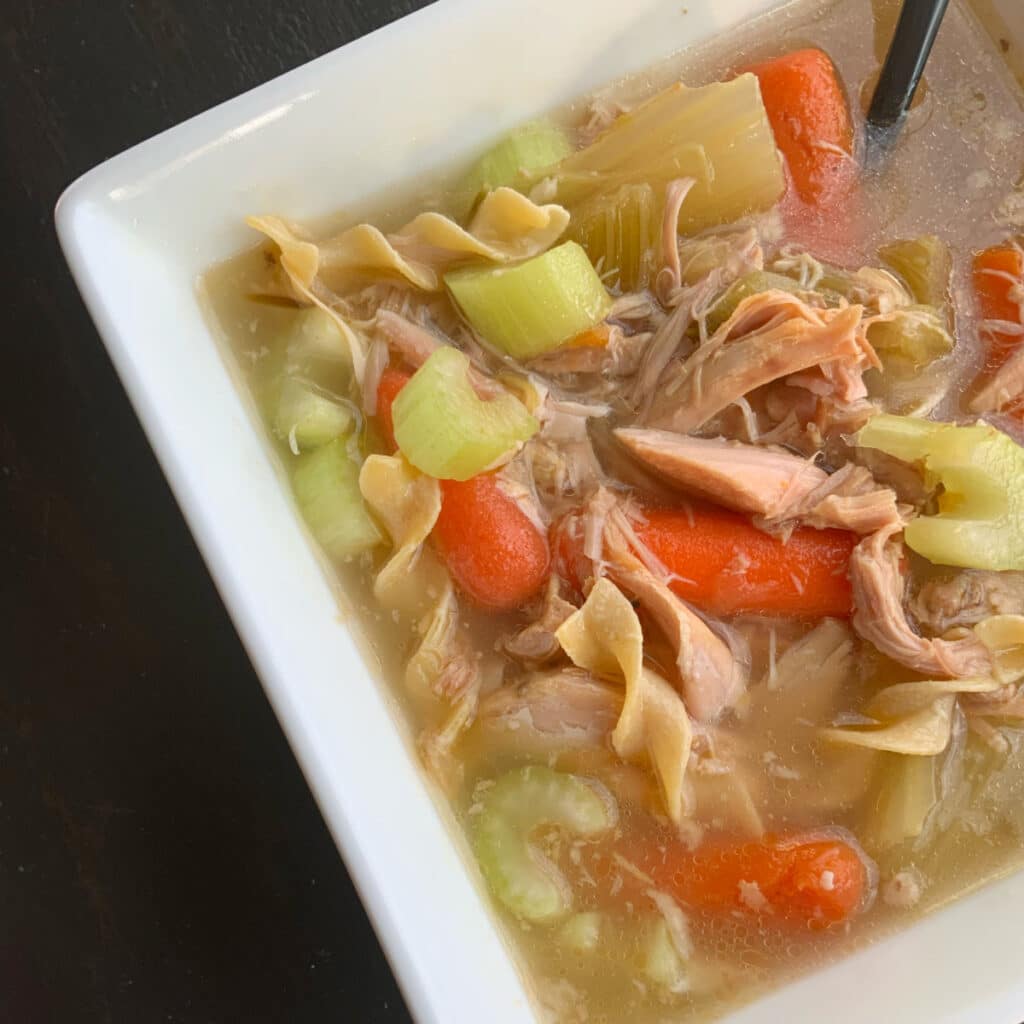 Turkey soup in a square bowl with a black spoon set on an angle. Soup has baby carrots, celery chunks and turkey shreds