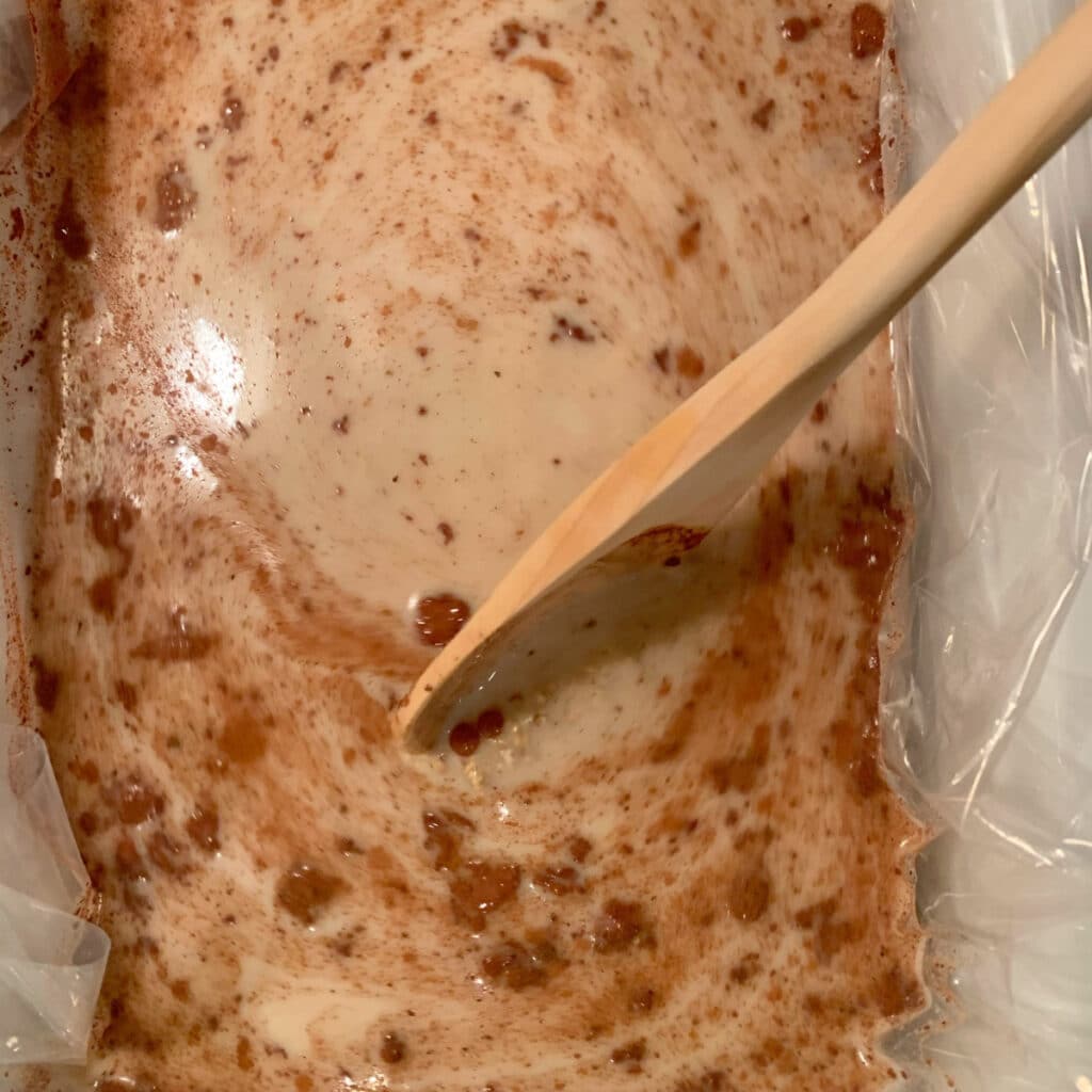 All raw ingredients in a white slow cooker vessel being stirred with a wooden spoon