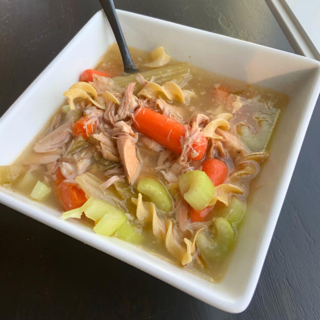 Close up of a bowl of turkey soup filled with carrots, celery, turkey and noodles. A black spoon is sticking out of the white square bowl that is holding the soup