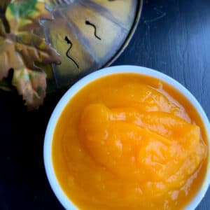 Close up of a round white bowl on a black table with bright orange purée in the bowl. A metal pumpkin is blurred off to the left side