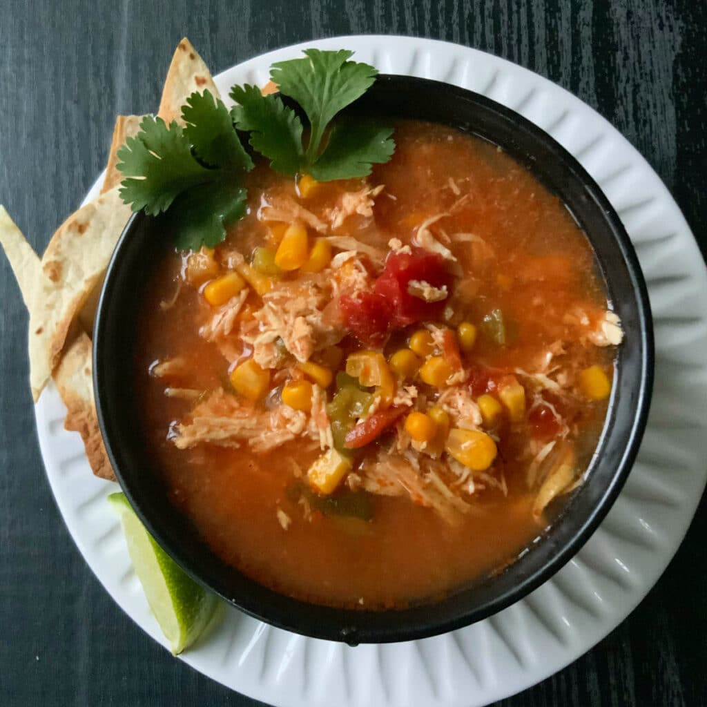 Looking down on a round black bowl that is on a round white plate filled with soup. Corn, shredded chicken and tomato chunks are visible.