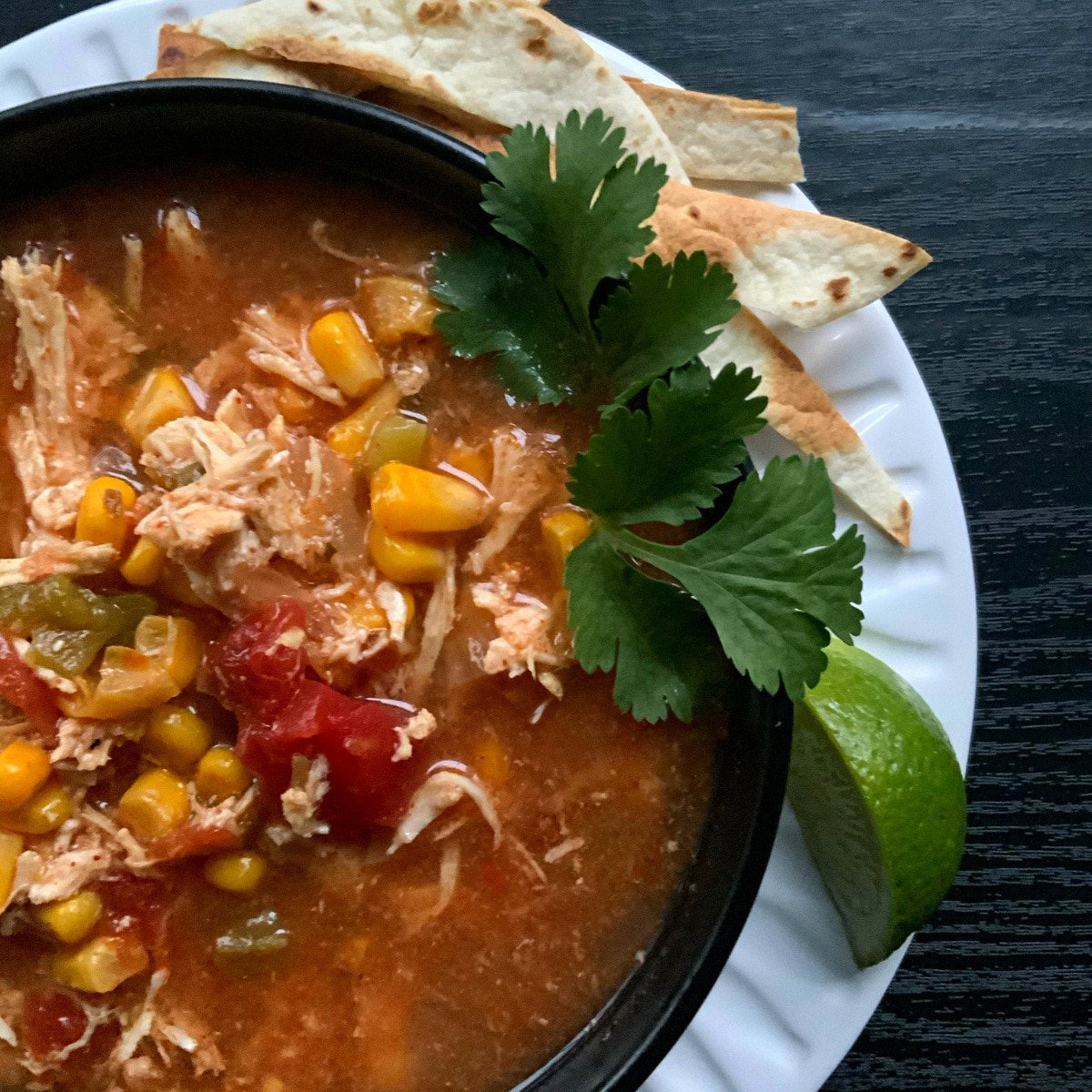 Chicken tortilla soup in a round black bowl garnished with green cilantro, a wedge of lime and some toasted tortilla strips