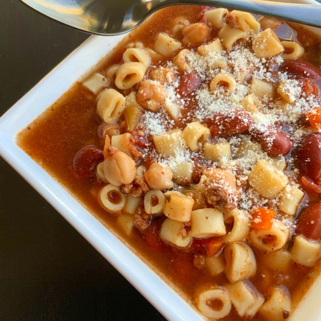 Close up side view of pasta e fagioli soup in a square white bowl with a dark spoon