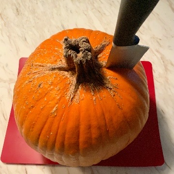 A whole, large pumpkin on a red cutting mat with a large knife stuck into it next to the stem