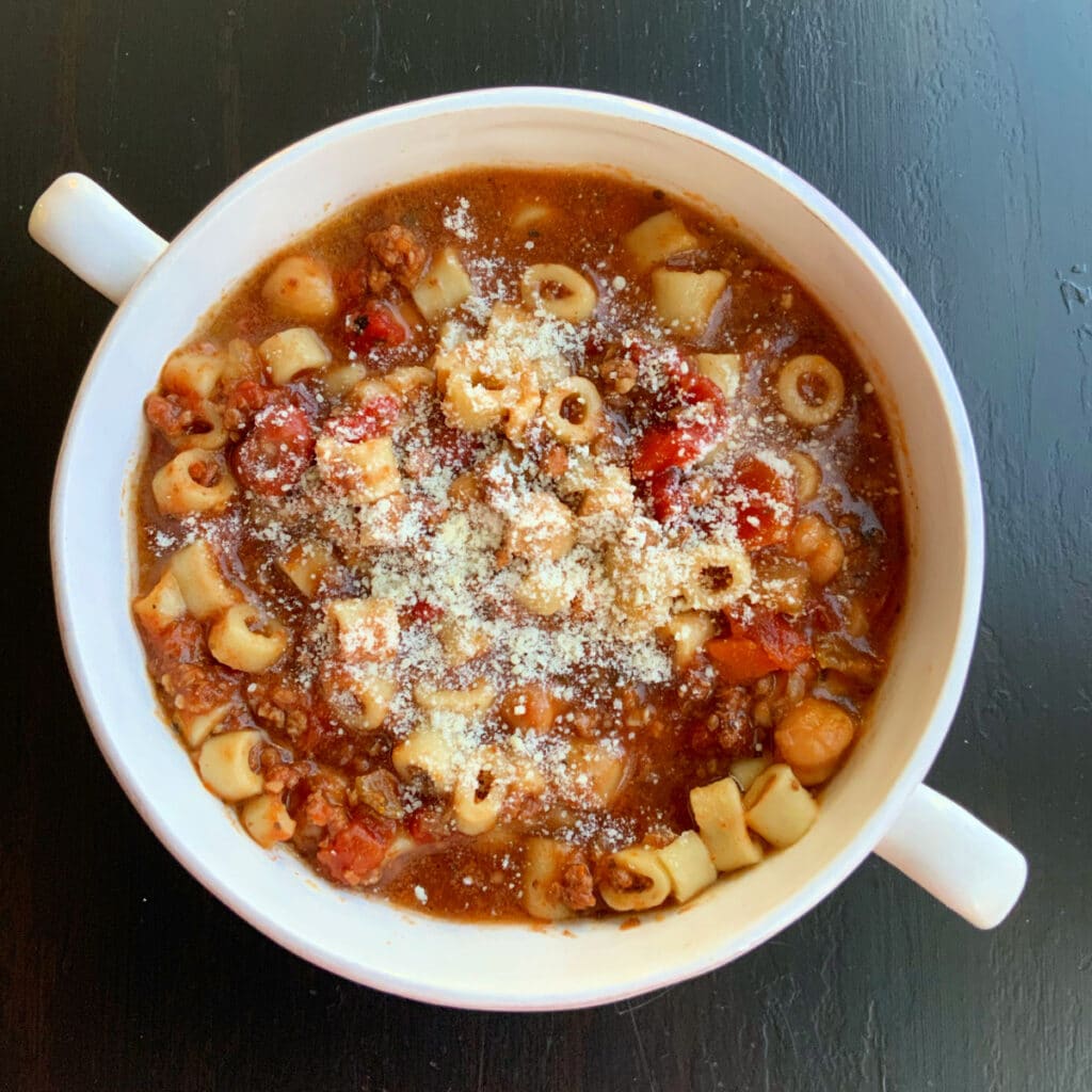 One round white bowl with two handles on a black table filled with soup, pasta, tomatoes and beans