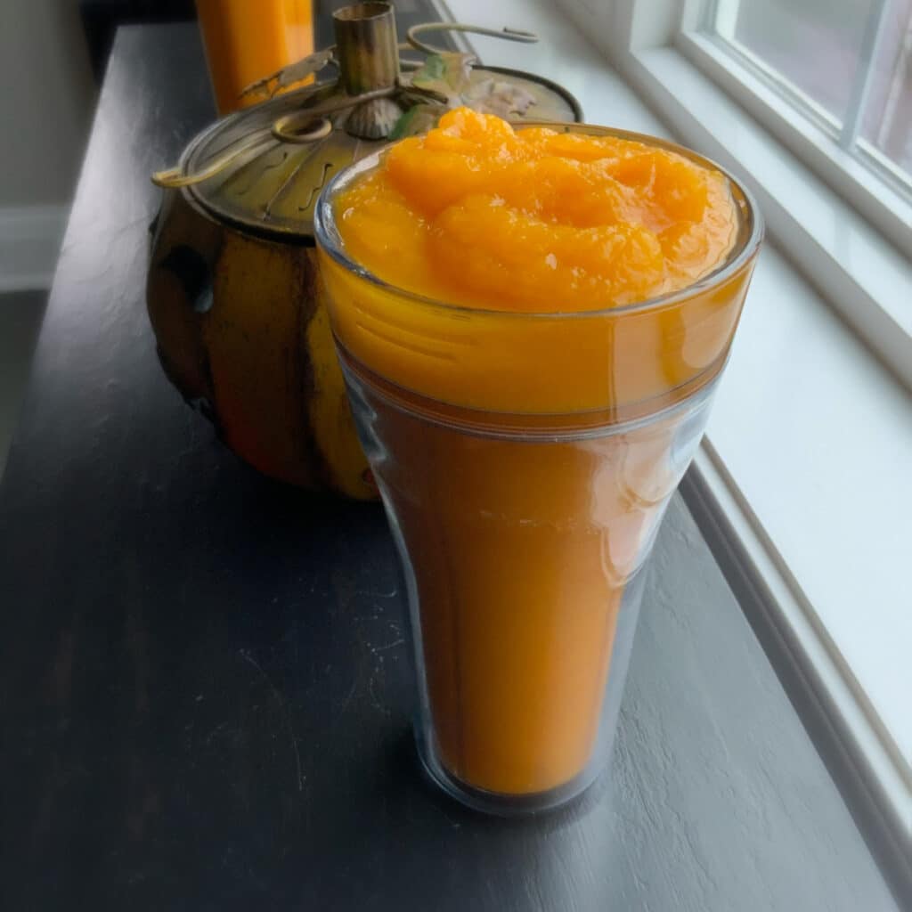 A tall plastic smoothie container filled with pumpkin purée sitting on a black table beside a window sill