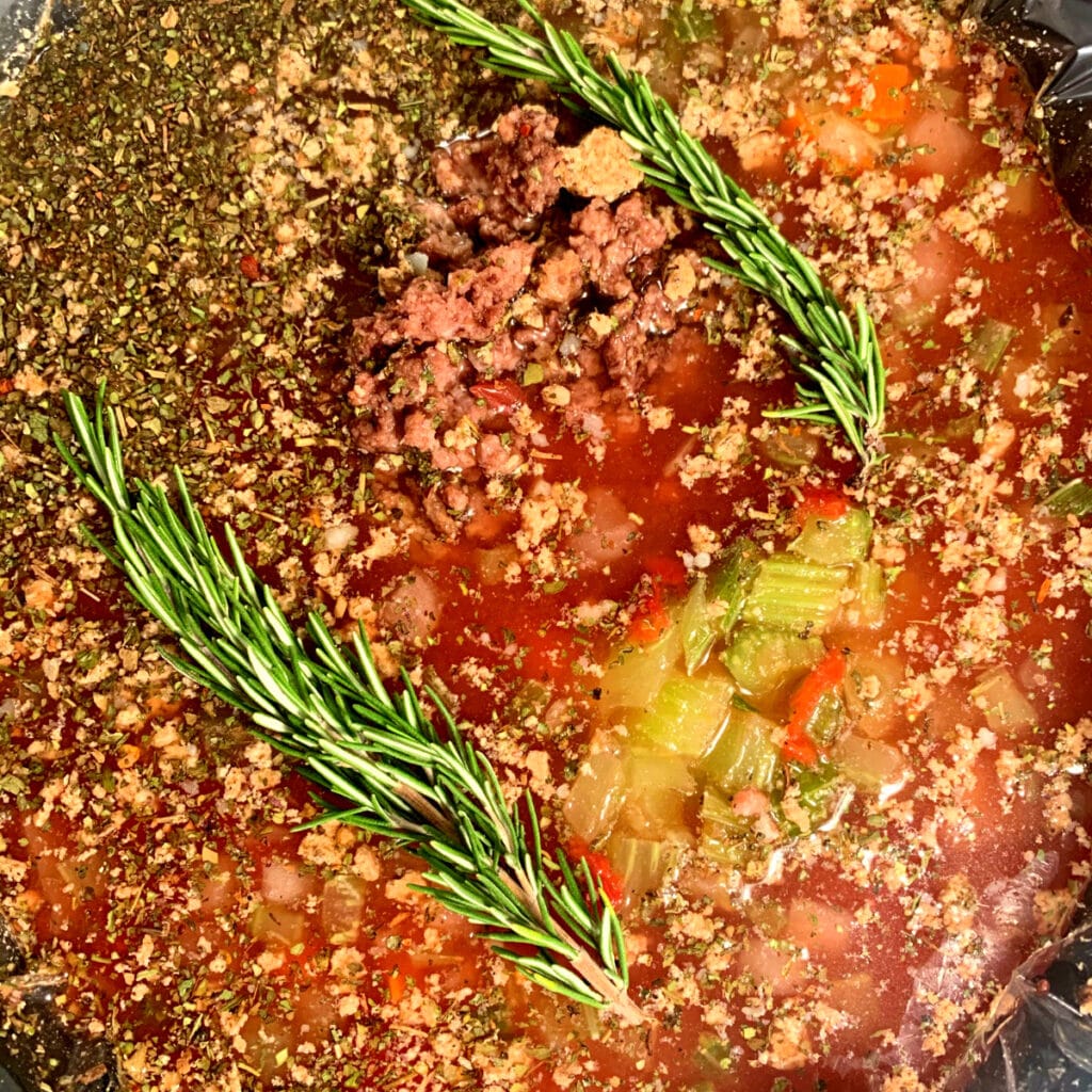 Close up of raw ingredients in slow cooker. Two fresh rosemary sprigs laying on top
