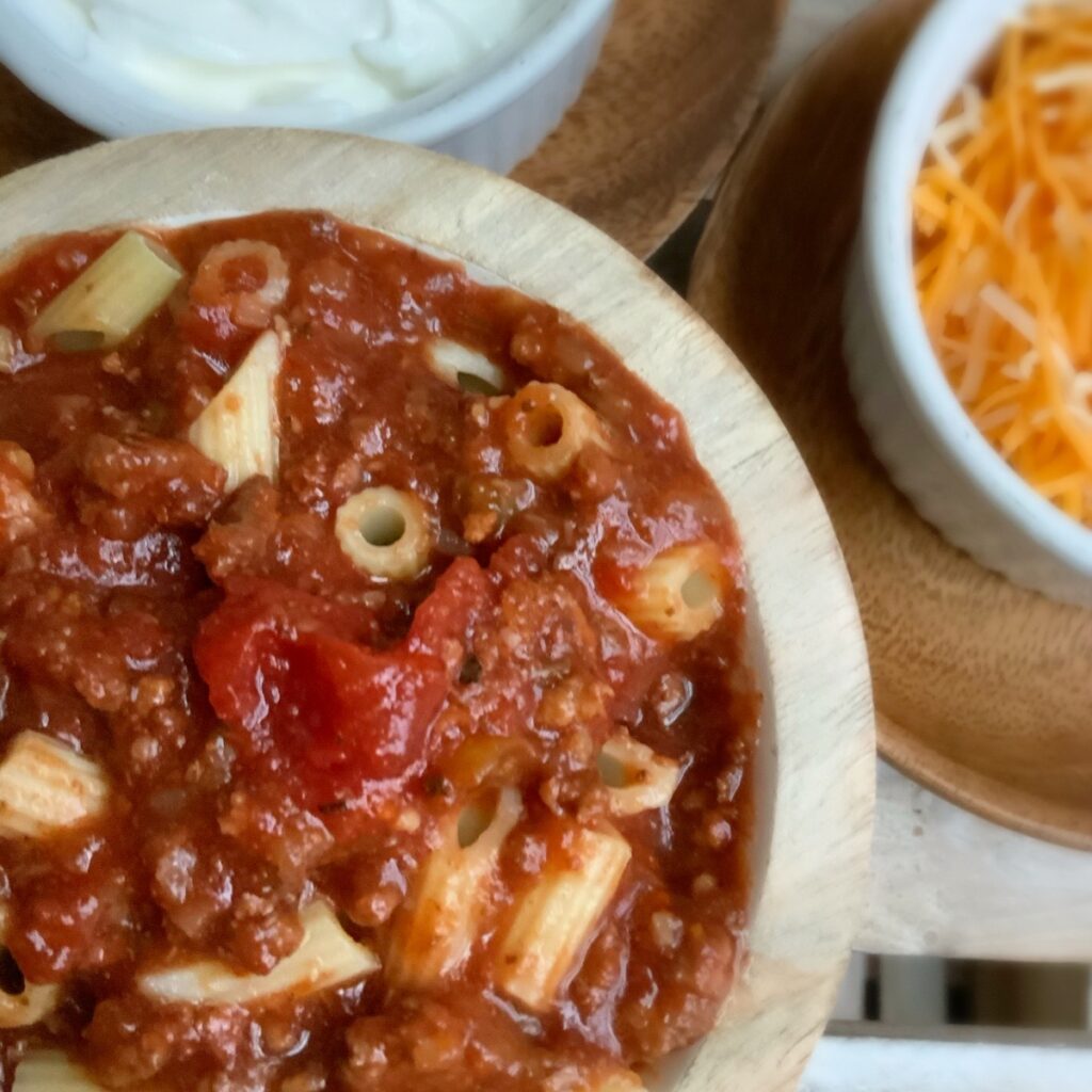 A round wooden bowl of goulash with a small round bowl of cheese and a small round bowl of sour cream