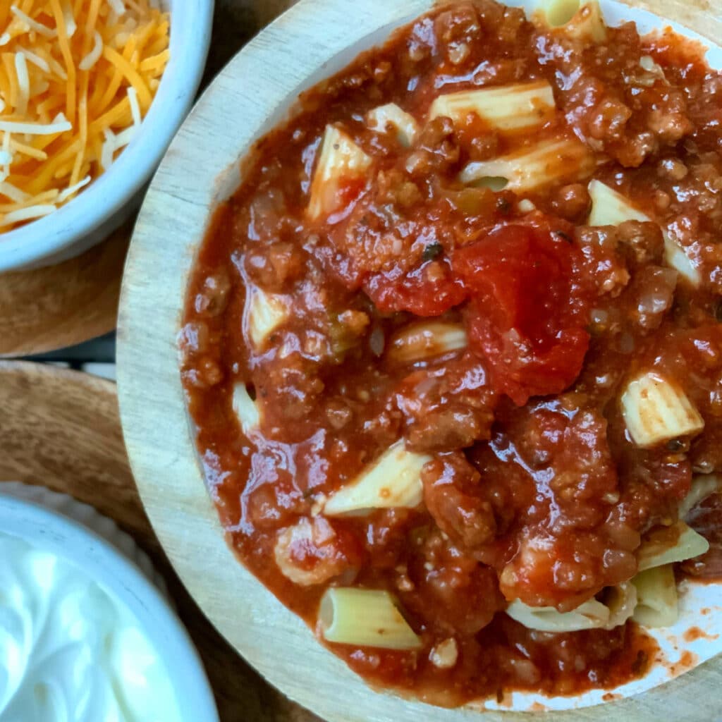A round red bowl filled with a beef and pasta dish in a tomato sauce flocked by a small bowl of shredded cheese and a small bowl of sour cream