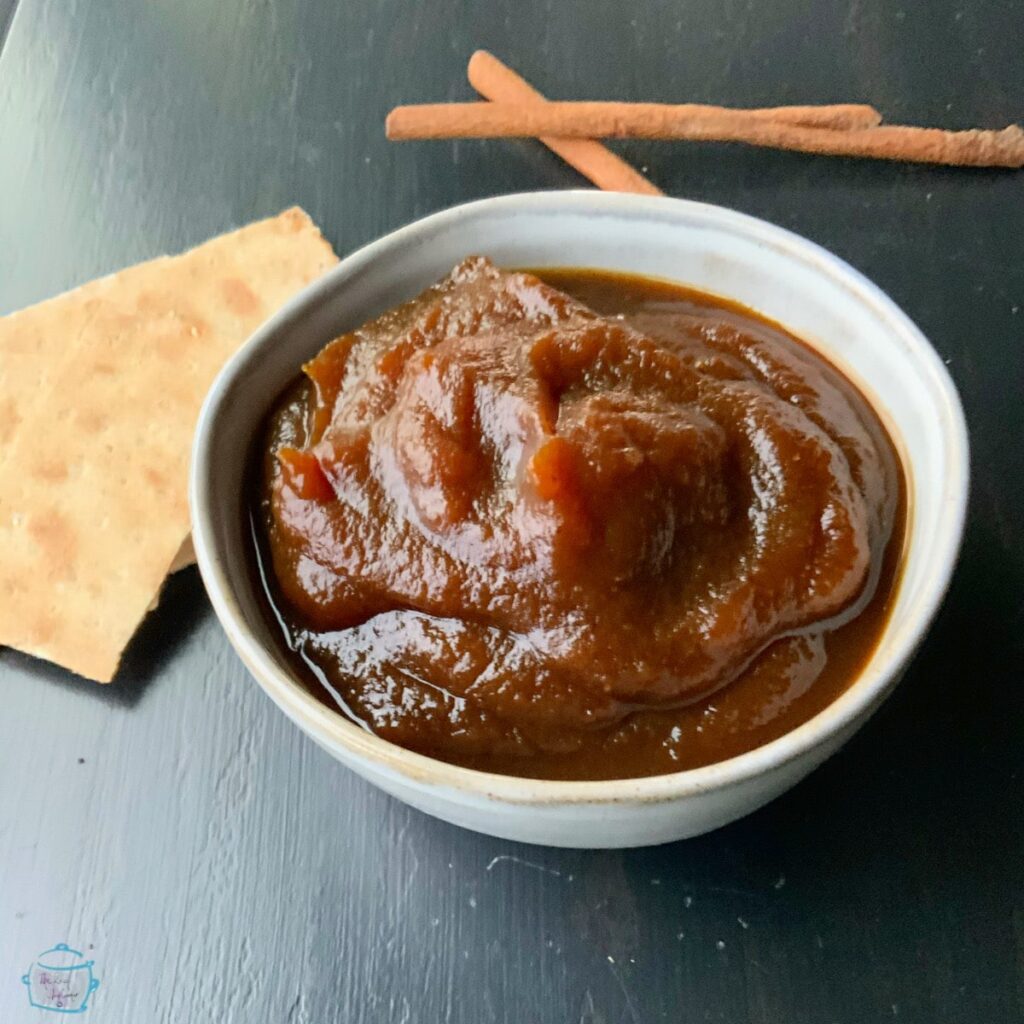 A side view of a grey bowl of spread with cinnamon sticks and crackers in background