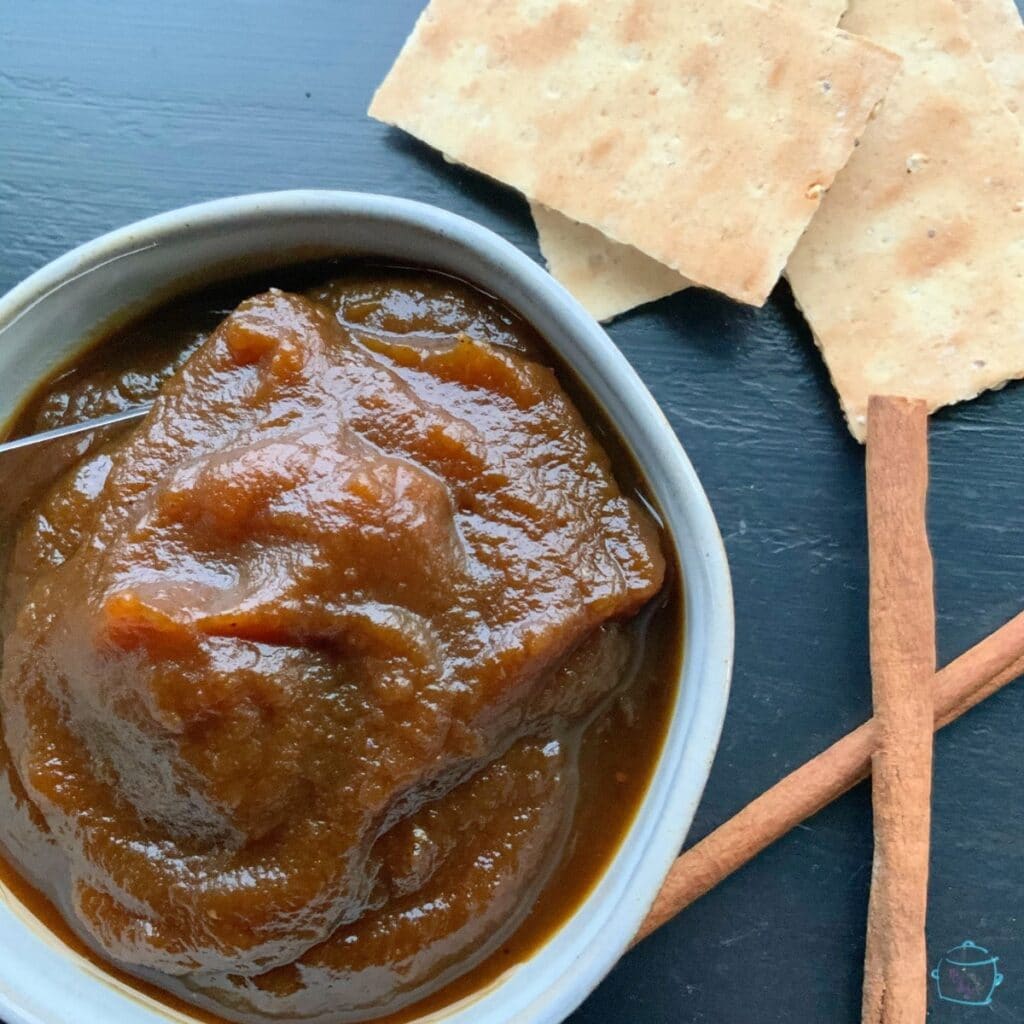 Gray bowl full of pumpkin butter that is ready to eat. Cinnamon sticks and crackers in background.