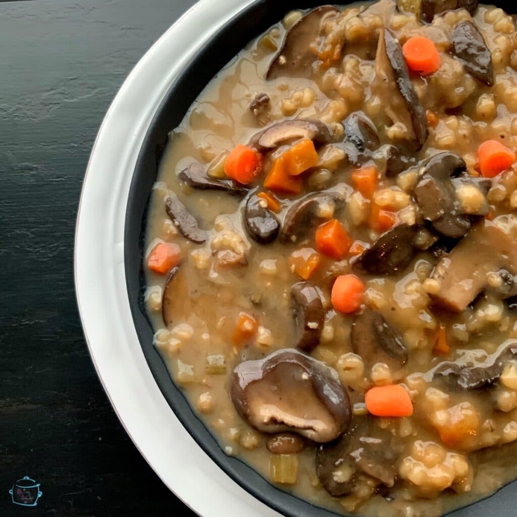 A round black bowl of thick soup filled with mushrooms, barley and veggies on a a white plate