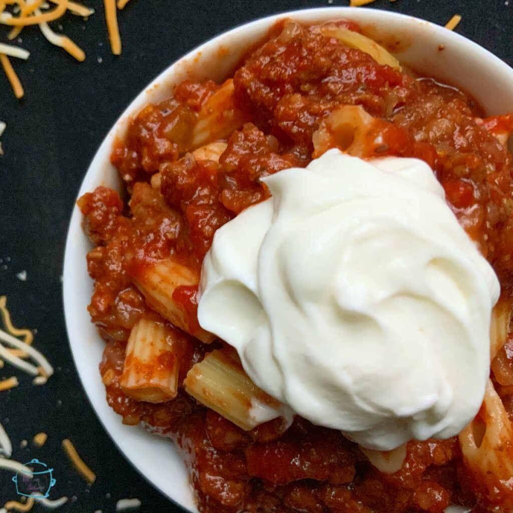 Close view of slow cooker goulash in a round white bowl sour cream on top and some shredded cheese laying around