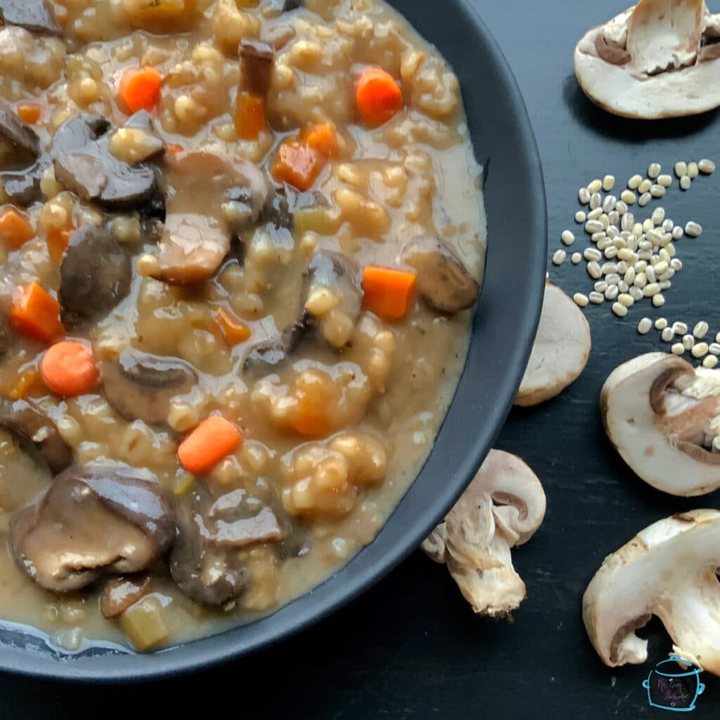 A round black bowl of mushroom barley soup with some raw mushrooms sliced and barley grains laying around it