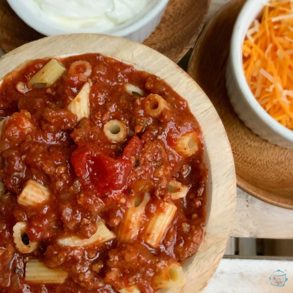 Slow cooker goulash in a round wooden bowl with smaller bowls of sour cream and shredded cheese blurred in the background