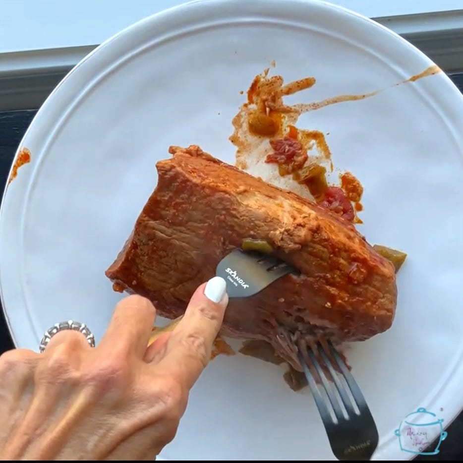 A cooked roast on a white plate with two hands holding two different forks beginning the shredding process
