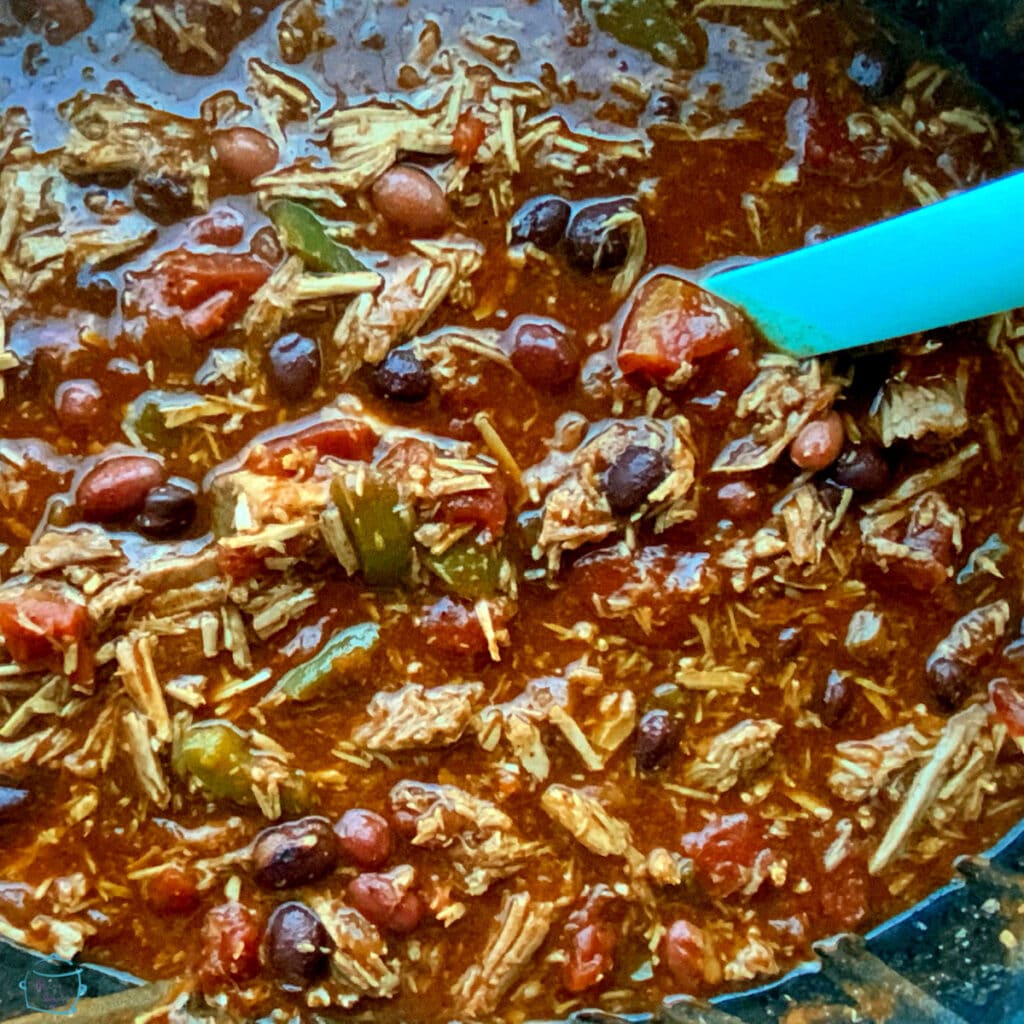 Close up of a crockpot filled with a saucy mix of shredded beef and beans being stirred by a large blue spoon