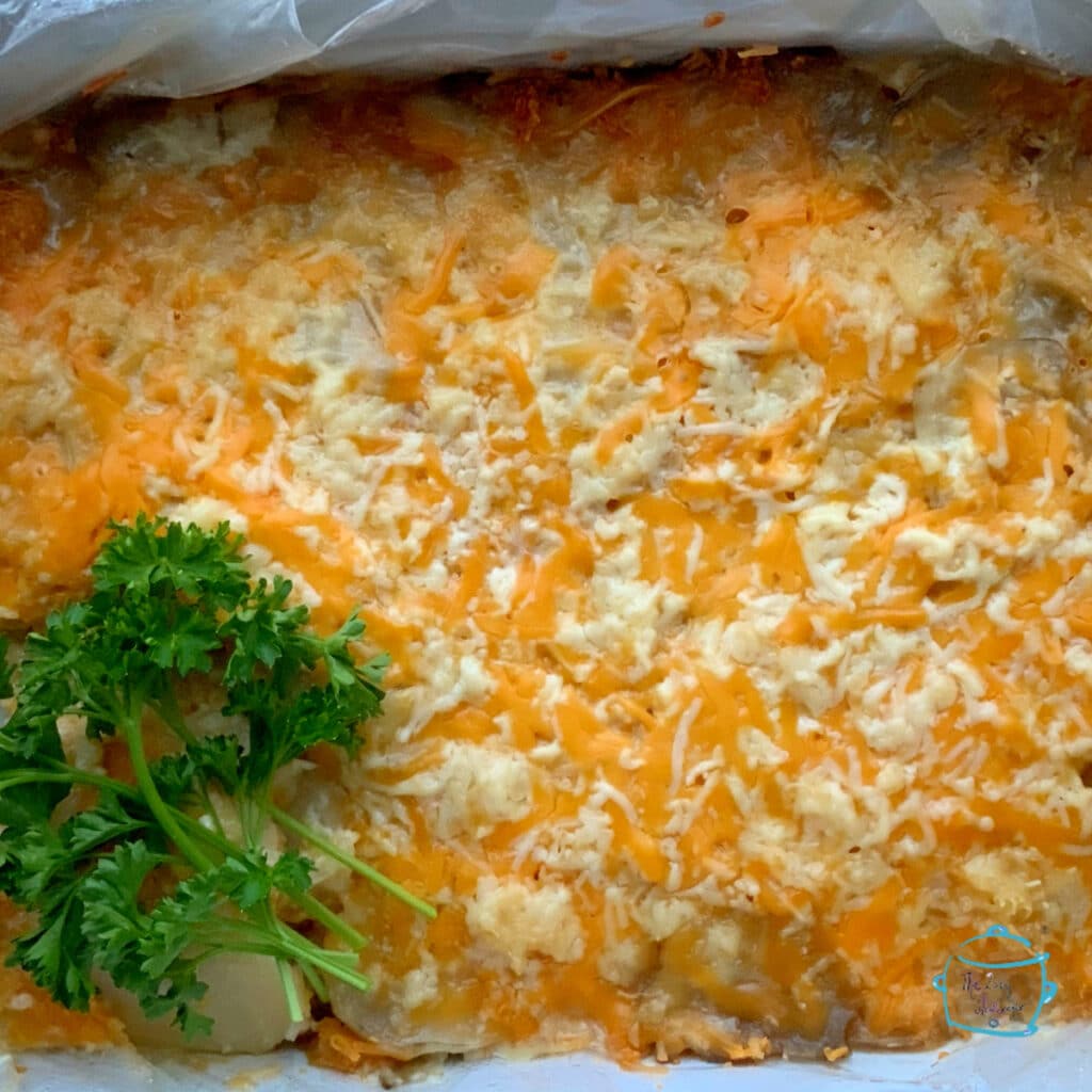 A full casserole shaped crockpot of cheesy potatoes with a white and orange cheese shreds melted on top and garnished with parsley off to the side