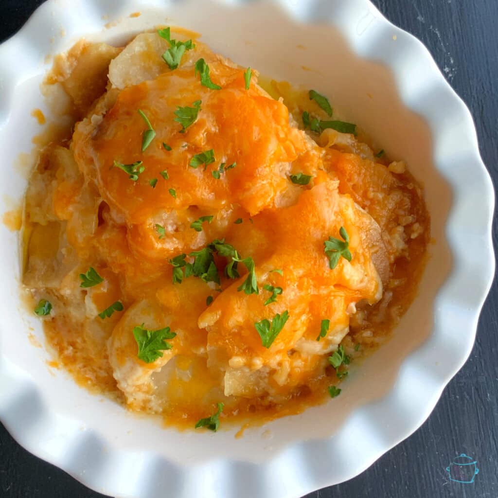 Looking down on a white bowl with wavy edged filled with potato slices, melted cheese and some chopped parsley for garnish