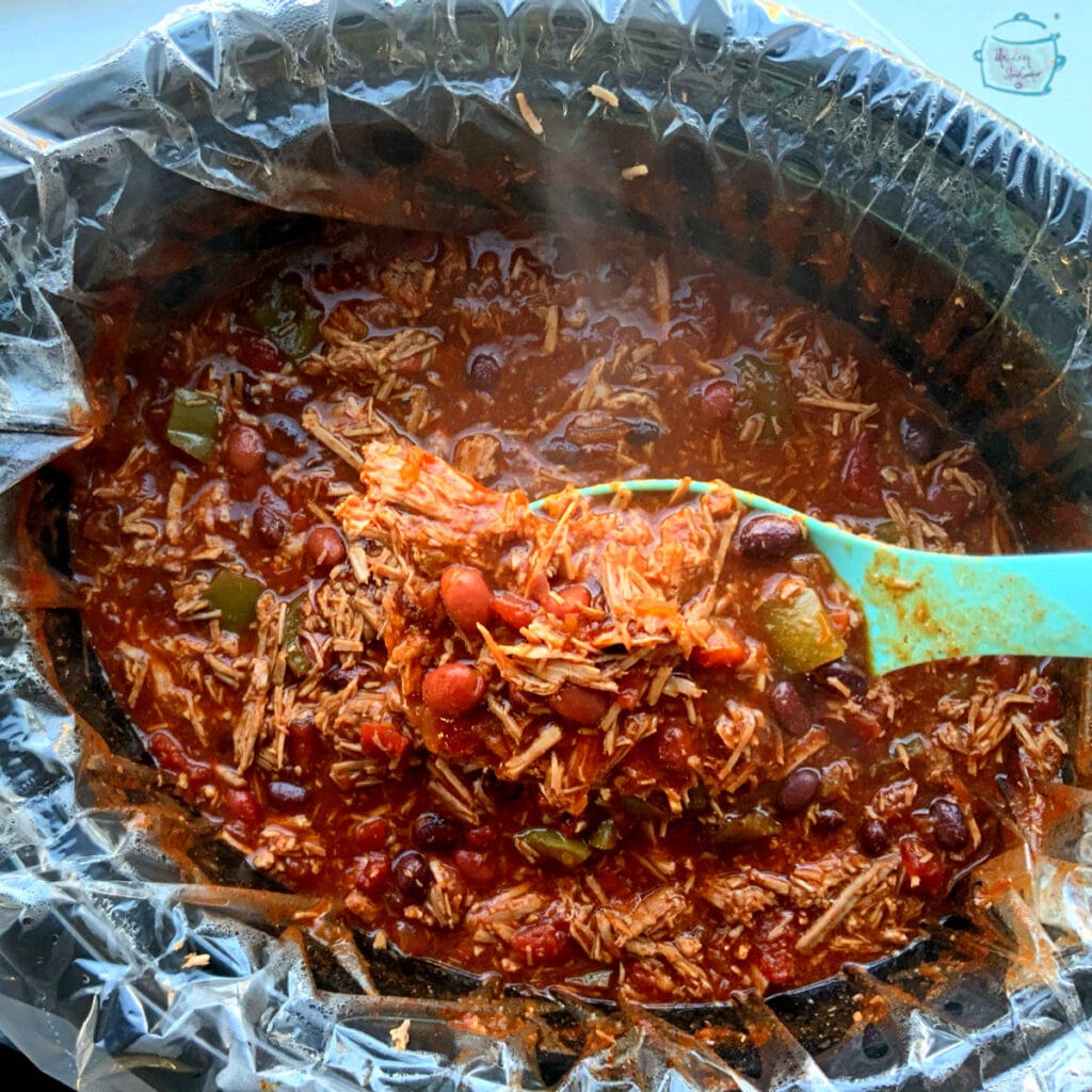 A slow cooker pot with a liner with chili in it, some chili is help closer to the camera with a large, turquoise spoon