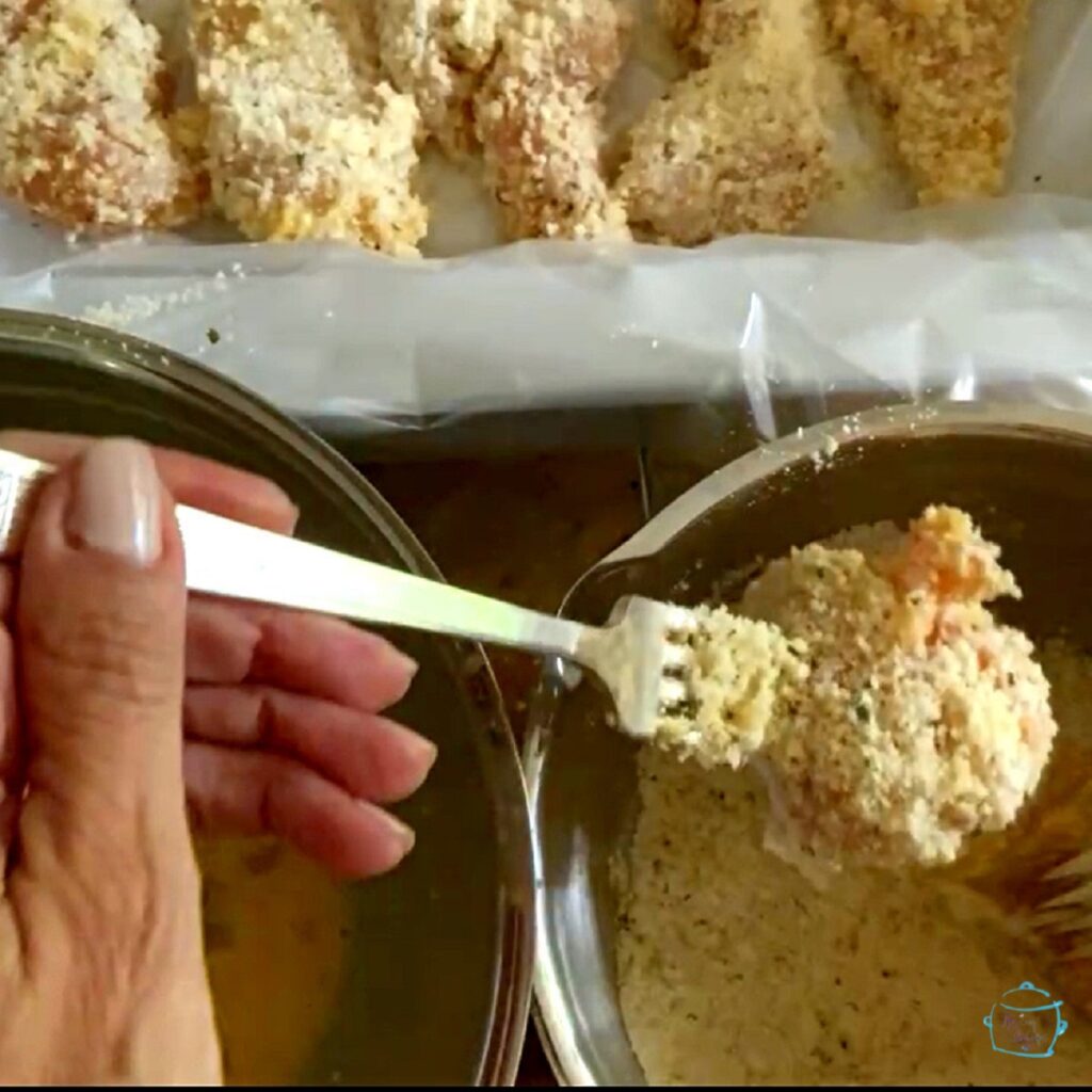 two bowls, one with eggs and one with bread crumbs a fork is holded is holding a piece of raw, breaded chicken over the bowls