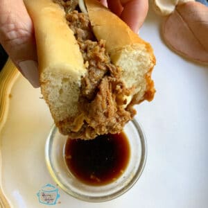 A hand holding a French dip sandwich that was just dipped in a clear bowl of au jus