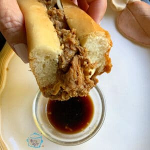 a roll filled with shredded beef held over a small glass bowl filled with au just ready to dip