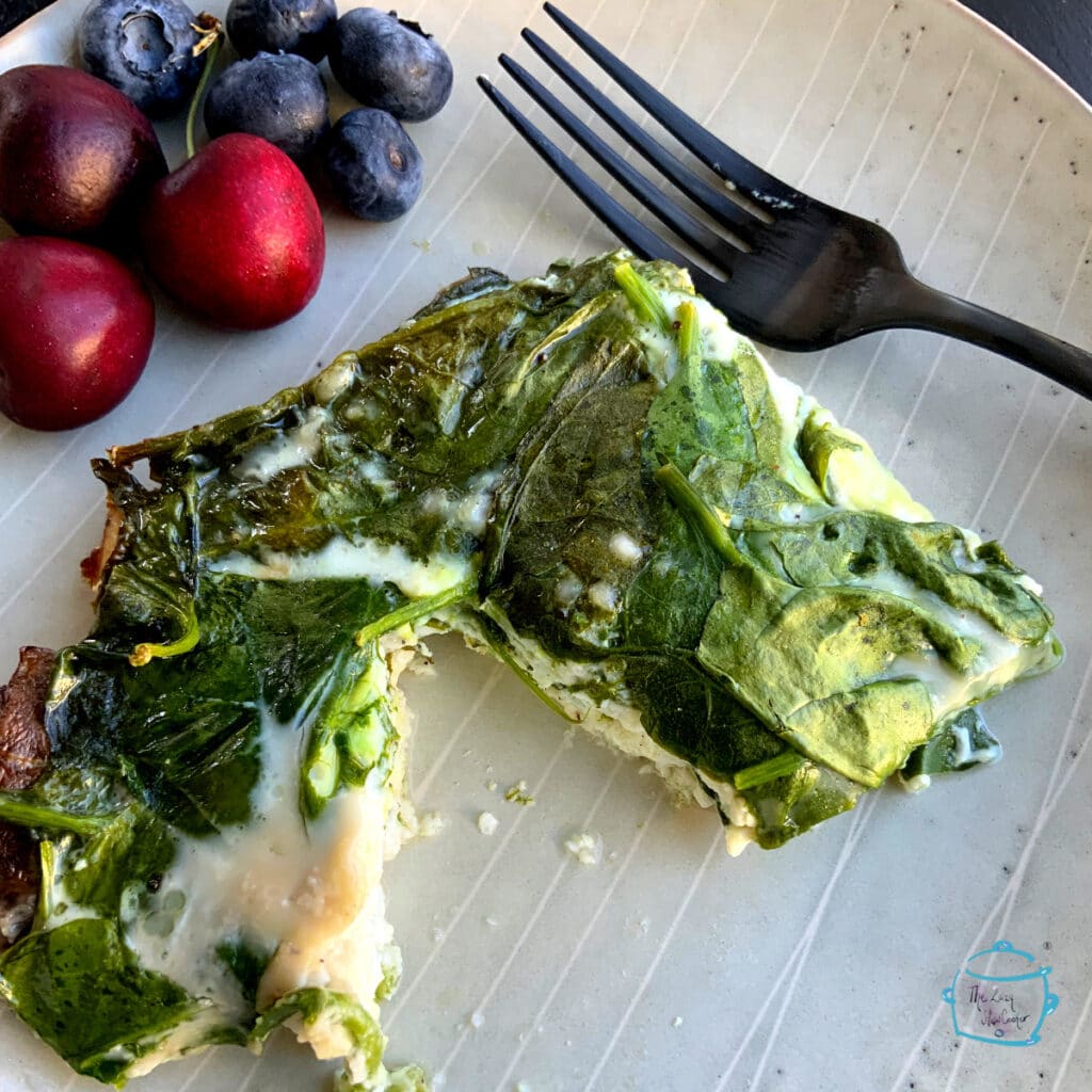 A piece of frittata on a plate with a fork and some fruit in the background