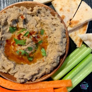 A round plate of baba ghanoush with carrots, celery and pita on the side