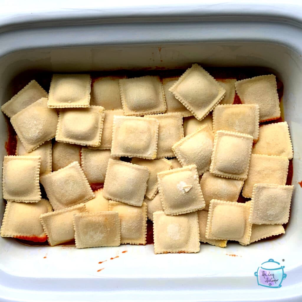 A casserole crockpot with a layer of frozen ravioli on top of red sauce