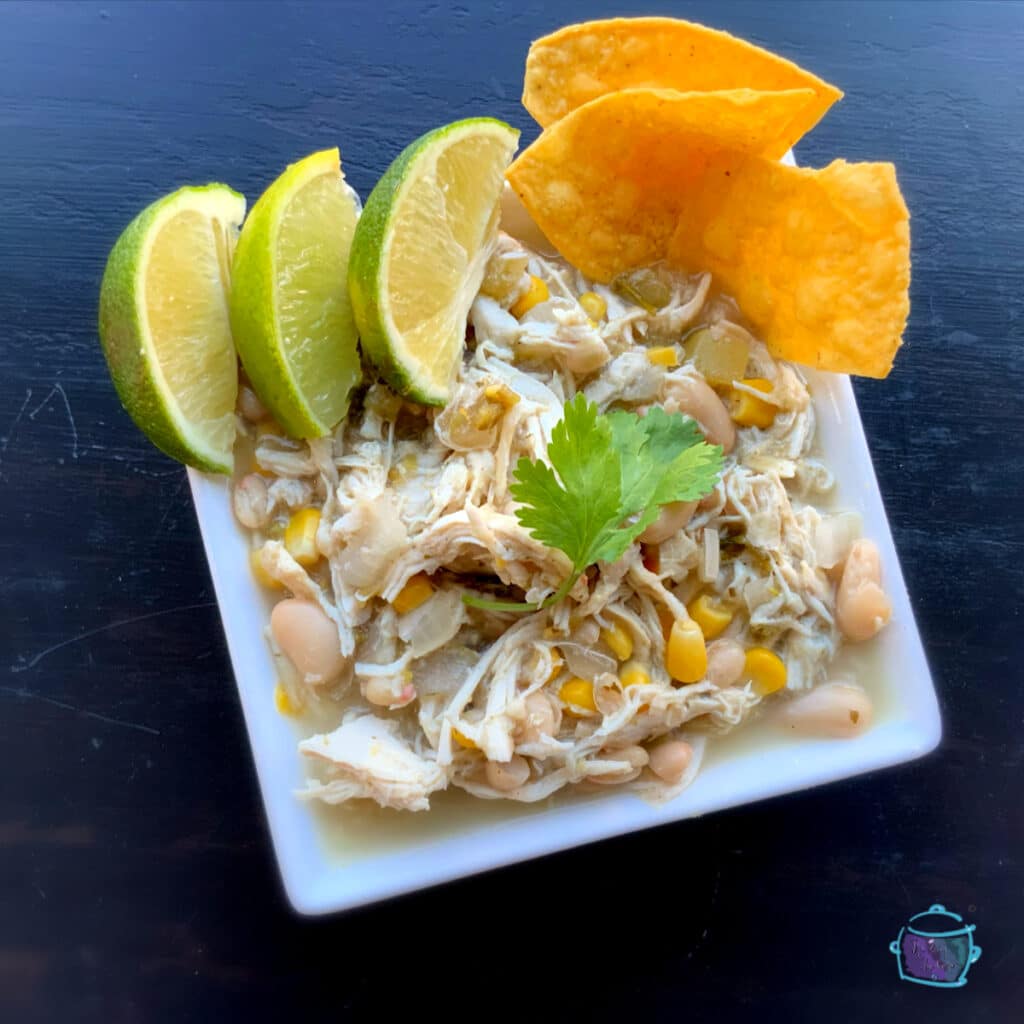 Looking down on a square bowl of chicken chili topped with a fresh cilantro leave with lime wedges and tortilla chips