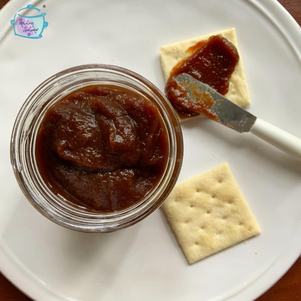 Apple butter in clear jar with some being spread on a nearby cracker