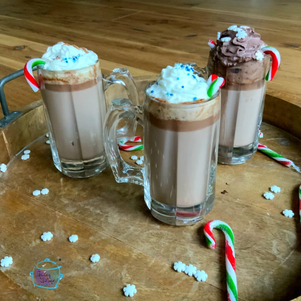 Three mugs of slow cooker hot chocolate topped with whipped cream and sprinkles