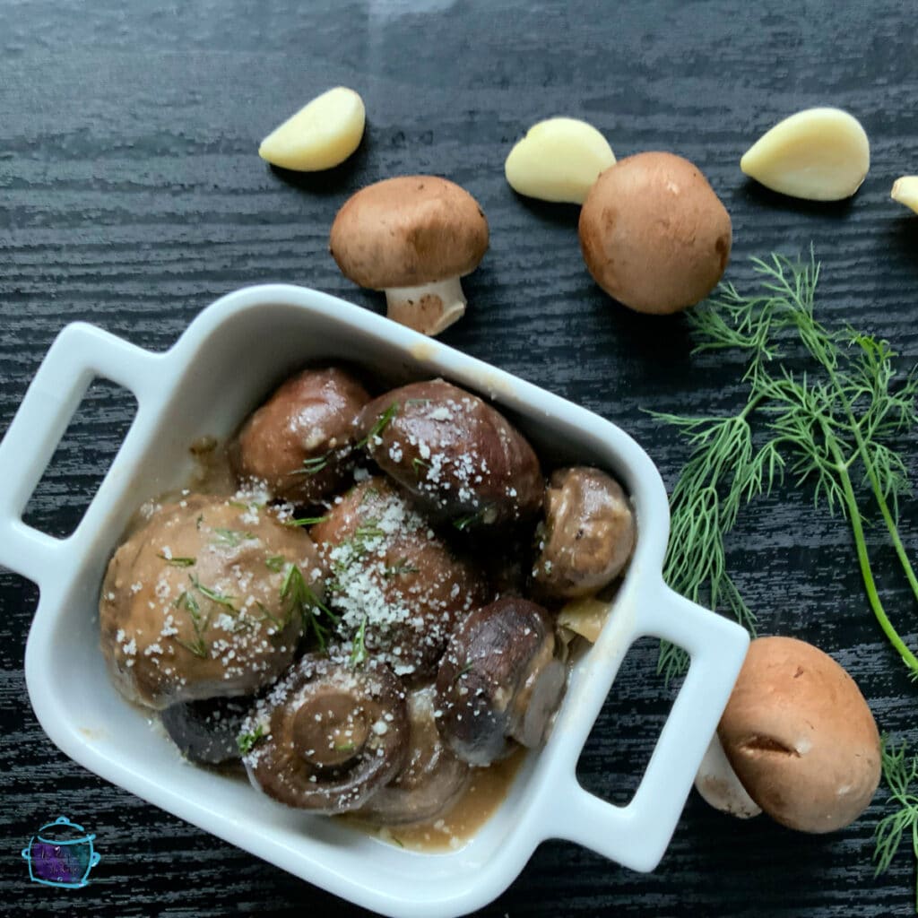 A square bowl containing finished garlic parmesan mushroom dish with some raw mushrooms garlic and herbs laying around