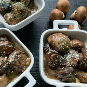 A square bowl containing finished garlic parmesan mushroom dish with some raw mushrooms garlic and herbs laying around