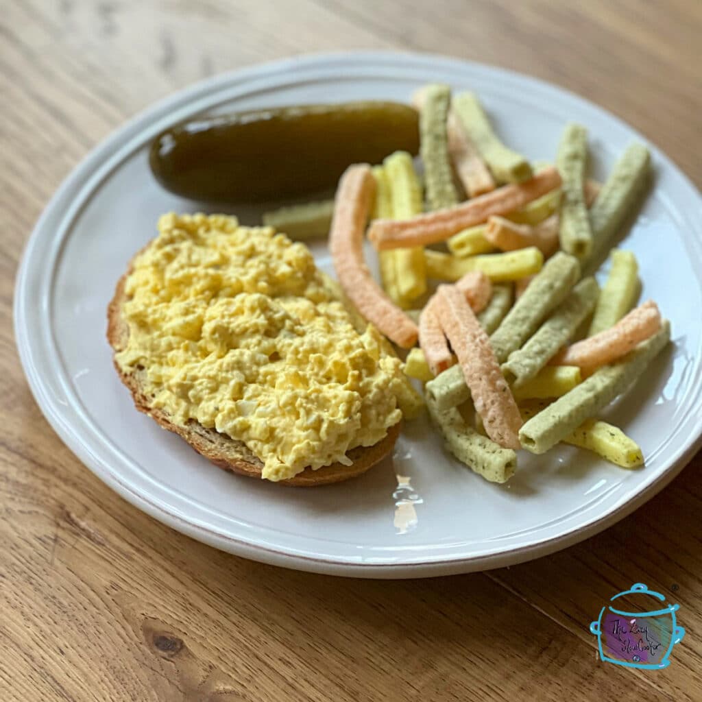 open faced egg salad sandwich on a plate with a pickle and chips