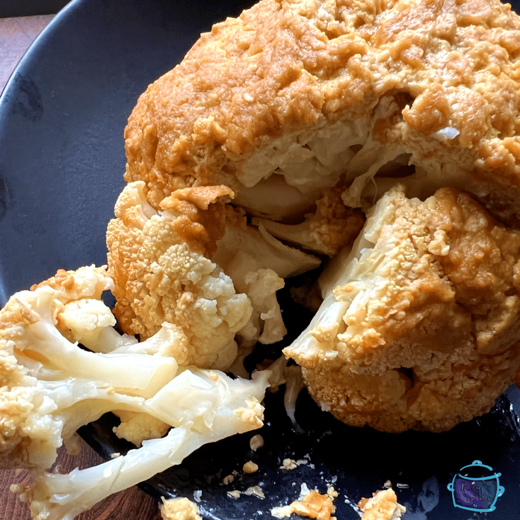 A full cooked head of cauliflower dijon parmesan on a black plate with a chunk cut out of it.