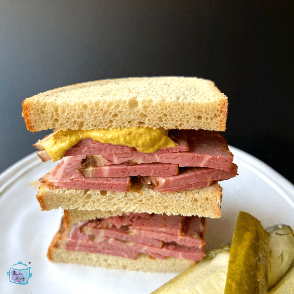 Pastrami sandwich halves piled on top of each other with mustard and a pickle