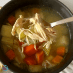 A bowl of chicken soup with a spoon in front of it full of shredded chicken, celery and broth