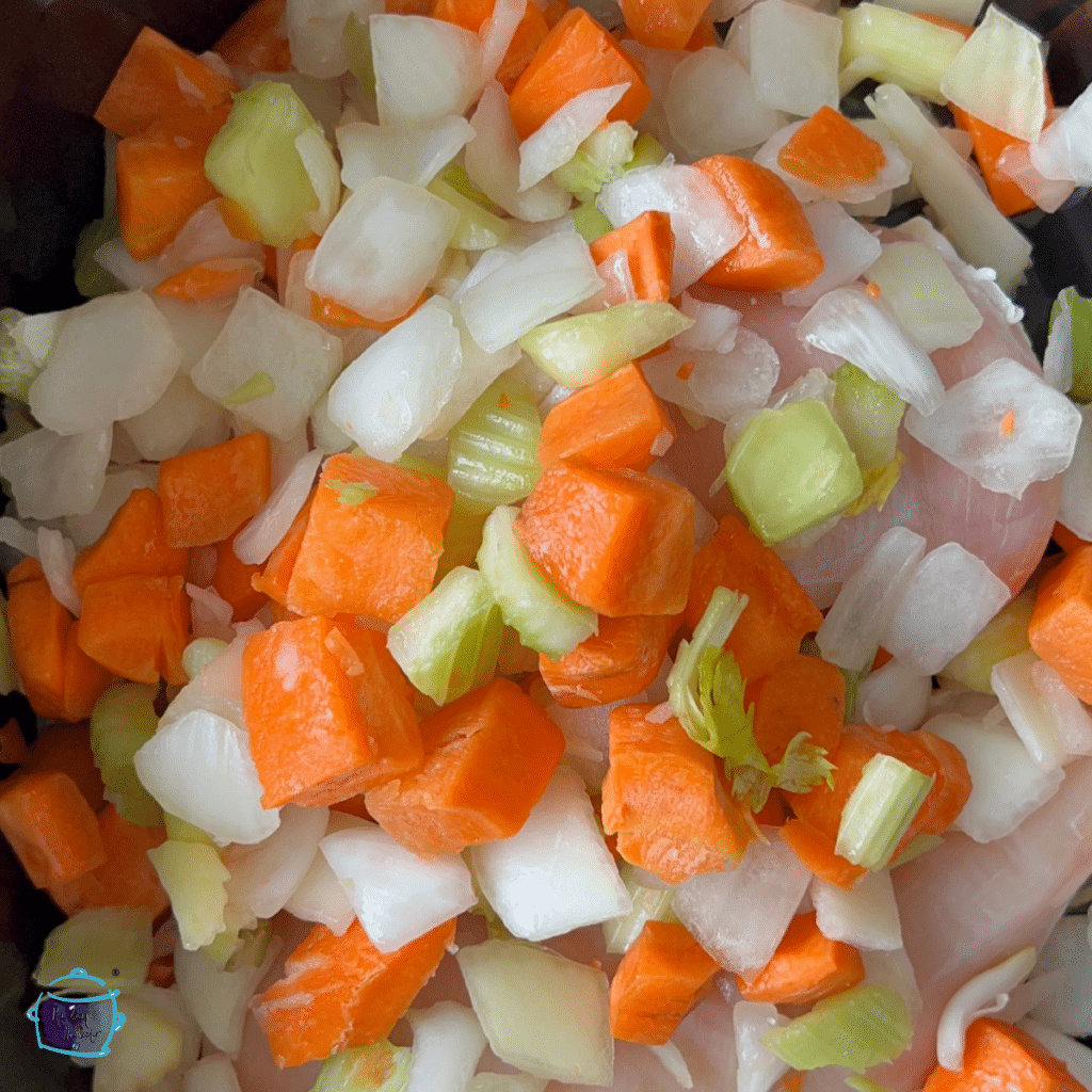 Close up of veggies cut up in the slow cooker on top of raw chicken pieces