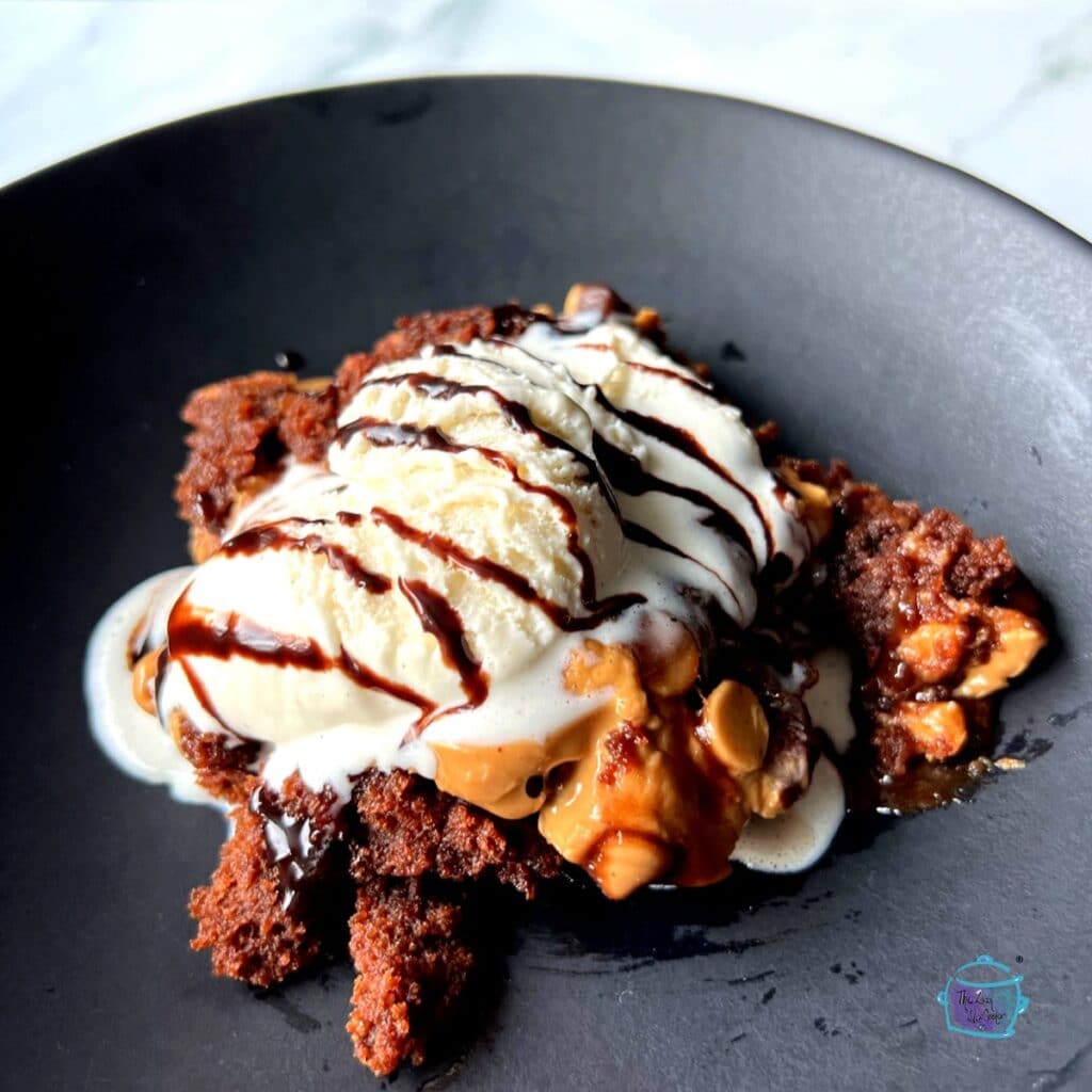 Peanut butter brownie on a black plate topped with vanilla ice cream and drizzled with chocolate fudge